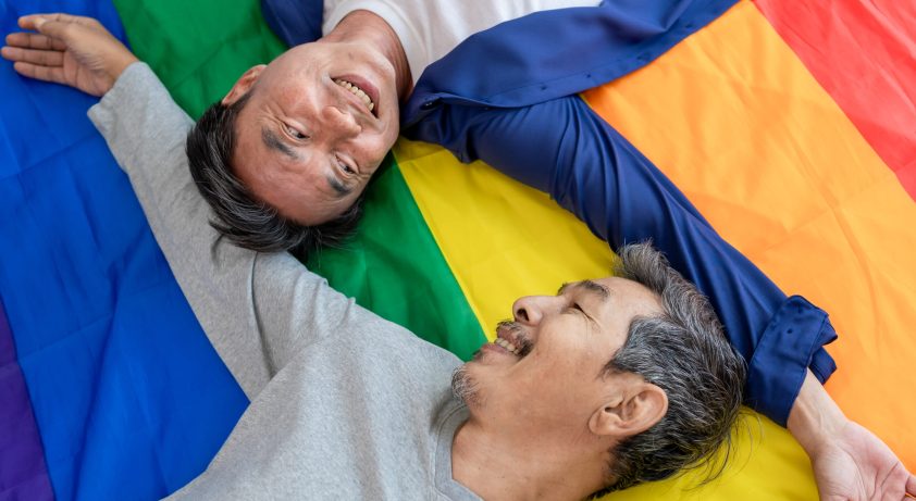 Asian senior elderly male gay couple lying down on bed with rainbow flag. Attractive mature man LGBTQ people looking at each other while spend morning routine after wake up together in bedroom at home